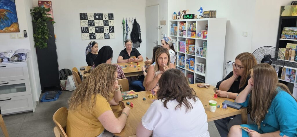 משתתפות משחקות במשחקי קופסה סביב שני שולחנות עץ, לצד מדפים מלאים במשחקים