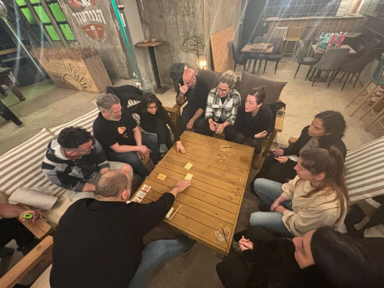 קבוצת מבוגרים יושבת סביב שולחן עץ ומשחקת במשחקי קופסה בסדנת גיבוש של משחקים מחוץ לקופסה